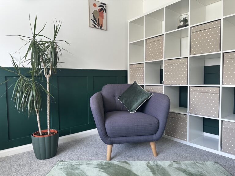 A calm counselling room with a grey armchair, green cushion, tall plant, and soft natural light creating a grounding, welcoming atmosphere.