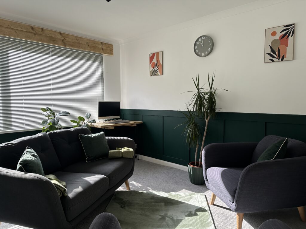 A modern therapy room featuring a grey sofa with green cushions, a desk by a bright window, abstract wall art, and indoor plants creating a warm, welcoming atmosphere.